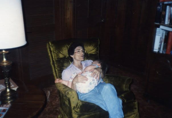 A snapshot from the 1980s: a woman in glasses sits in a green velvet armchair, holding a sleeping baby against her chest, a brass lamp and bookshelves behind her.